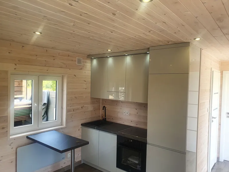 Kompakte Einbauküche mit hellgrauen Hochglanzfronten, dunkler Arbeitsplatte und integrierten LED-Spots an der Holzdecke sowie einem ausklappbaren Tisch in einem Bungalow der MONO-Serie.