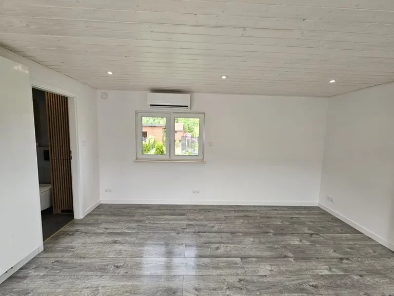 Geräumiger Wohnbereich im modernen skandinavischen Stil in einem winterfesten Bungalow der MONO-Serie von Compact Haus mit Klimaanlage, Holzoptikboden und weiß lasierter Decke.