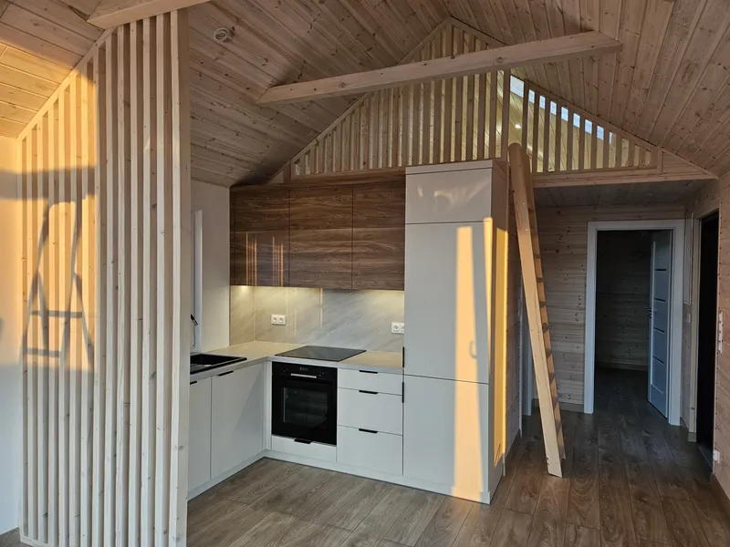 Moderne Einbauküche im Loft-Stil eines Bungalows der MONO-Serie von Compact Haus mit einer Trennwand aus Holzlamellen, einer Holzleiter und gemütlichem Sonnenlicht.