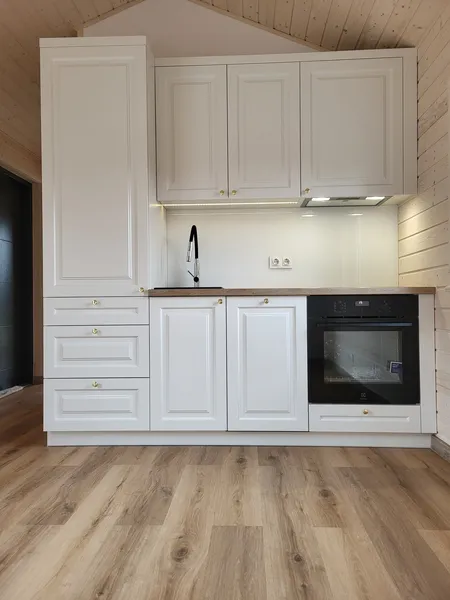 Kompakte weiße Küchenzeile im Landhausstil mit integriertem Backofen, schwarzer Armatur, heller Glasrückwand und Holzoptik-Boden in einem Bungalow der MONO-Serie.