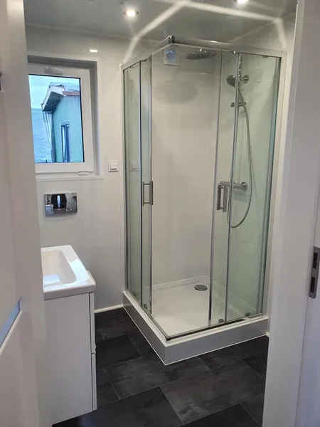 Helles Badezimmer in einem schlüsselfertigen Ferienhaus der MONO-Serie von Compact Haus mit Eck-Glasdusche, Regendusche und Fliesen in Schieferoptik.