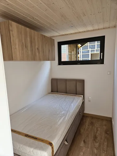 Kompakter Schlafbereich in einem Ferienhaus der MONO-Serie von Compact Haus mit Einzelbett, Hängeschrank in Holzoptik und anthrazitfarbenem Fenster.
