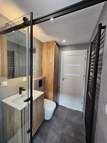 Modernes Badezimmer in einem schlüsselfertigen Minihaus der MONO-Serie von Compact Haus mit barrierefreier Glasdusche, Holzmöbeln in Lamellenoptik und Handtuchheizkörper.