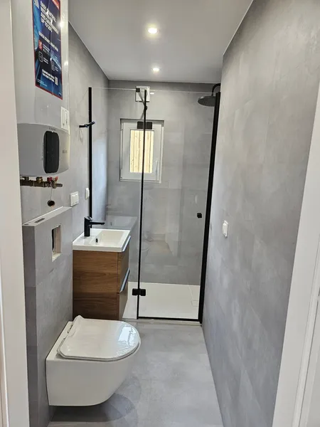 Modernes, schmal geschnittenes Badezimmer in einem schlüsselfertigen Minihaus der MONO-Serie von Compact Haus mit barrierefreier Glasdusche, Holzwaschtisch und großformatigen Fliesen.