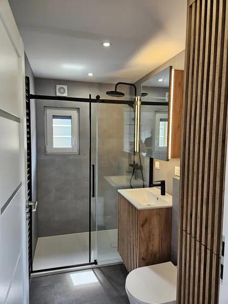 Luxus-Badezimmer in einem schlüsselfertigen Minihaus der MONO-Serie von Compact Haus mit bodengleicher Glasdusche, Holzwaschtisch in Lamellenoptik und beleuchtetem Spiegelschrank.