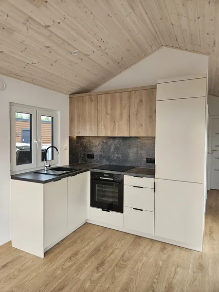 Kompakte L-Küche in einem Minihaus der MONO-Serie von Compact Haus mit cremeweißen Unterschränken, Oberschränken in Eichenoptik und einer dunklen Rückwand in Schieferoptik.