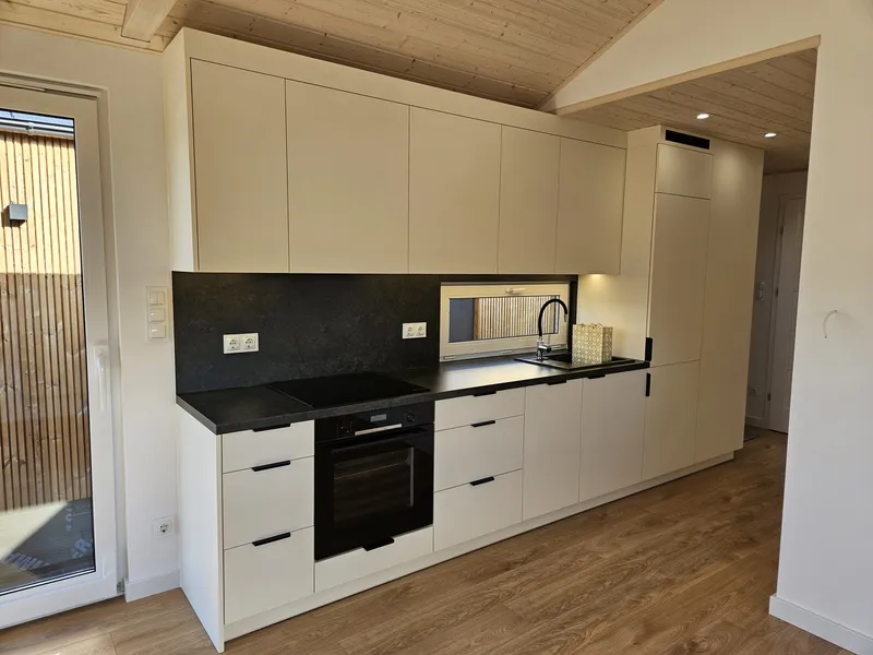 Moderne, lange Küchenzeile in Creme mit integriertem Backofen, Induktionskochfeld und einer kontrastierenden dunklen Rückwand im Innenraum eines Minihauses der MONO-Serie.
