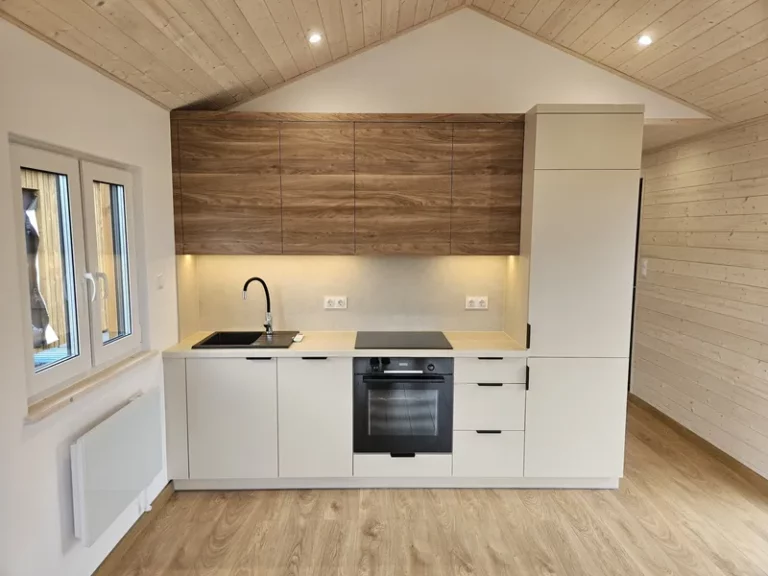 Moderne, grifflose Küchenzeile in einem Minihaus der MONO-Serie von Compact Haus mit cremeweißen Fronten, Oberschränken in dunkler Holzoptik und integriertem Backofen.