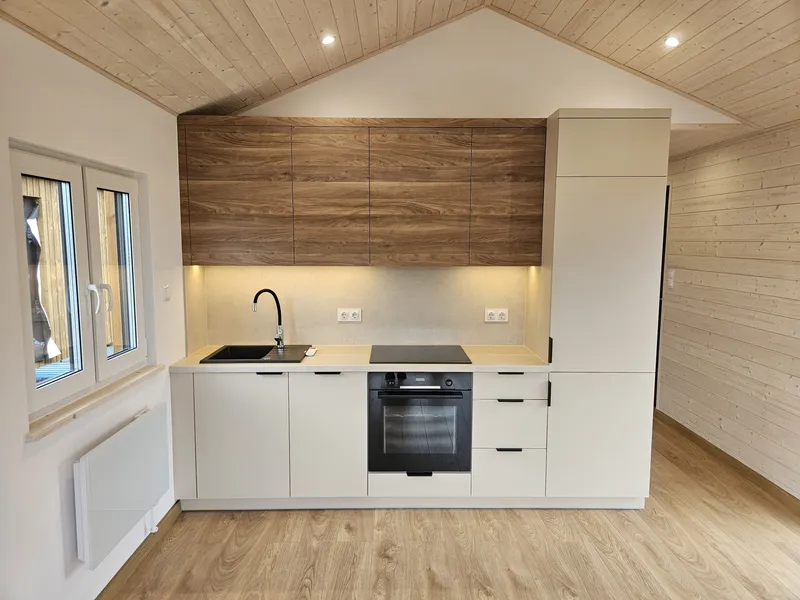 Moderne, grifflose Küchenzeile in einem Minihaus der MONO-Serie von Compact Haus mit cremeweißen Fronten, Oberschränken in dunkler Holzoptik und integriertem Backofen.