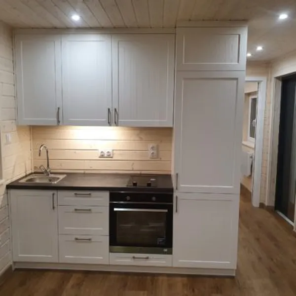 Kompakte weiße Küchenzeile im Landhausstil mit integriertem Backofen, Kochfeld und Spüle vor einer hellen Holzwand im Minihaus der MONO-Serie.
