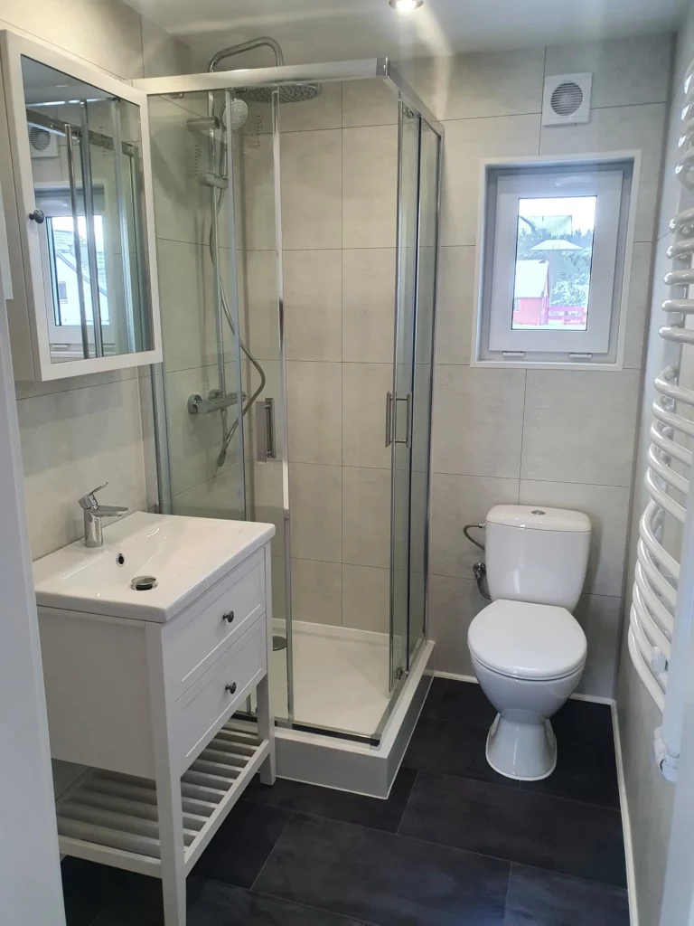 Helles Badezimmer in einem Mobilheim der MONO-Serie von Compact Haus mit einer gläsernen Eckdusche, weißem Waschtisch, Spiegelschrank und dunklem Fliesenboden.