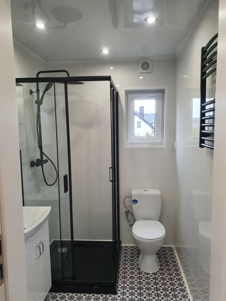 Badezimmer in einem Compact Haus der MONO-Serie mit maßgefertigtem Hochschrank aus Holzlamellen, Wand-WC, Spiegel und Blick durch die geöffnete Tür in das angrenzende Zimmer mit grauem Vinylboden.