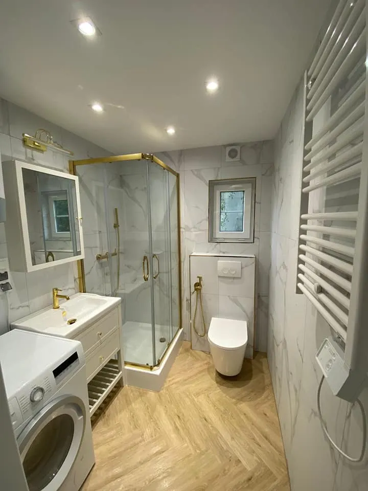 Badezimmer im Mobilheim der MONO-Serie von Compact Haus mit einer Waschmaschine, Marmoroptik-Fliesen und goldenen Armaturen an Dusche und Waschbecken.