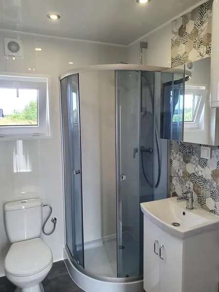 Modernes Badezimmer in einem winterfesten Mobilheim der MONO-Serie von Compact Haus mit runder Glasdusche, weißem Waschtisch, dekorativen Mosaikfliesen und natürlichem Tageslicht.