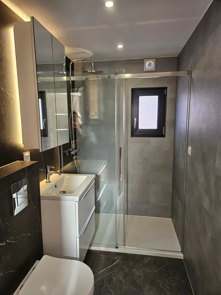 Luxuriöses Badezimmer in einem winterfesten Mobilheim der MONO-Serie von Compact Haus mit barrierefreier Glasdusche, Fliesen in Beton- und Marmoroptik sowie LED-Spiegelschrank.