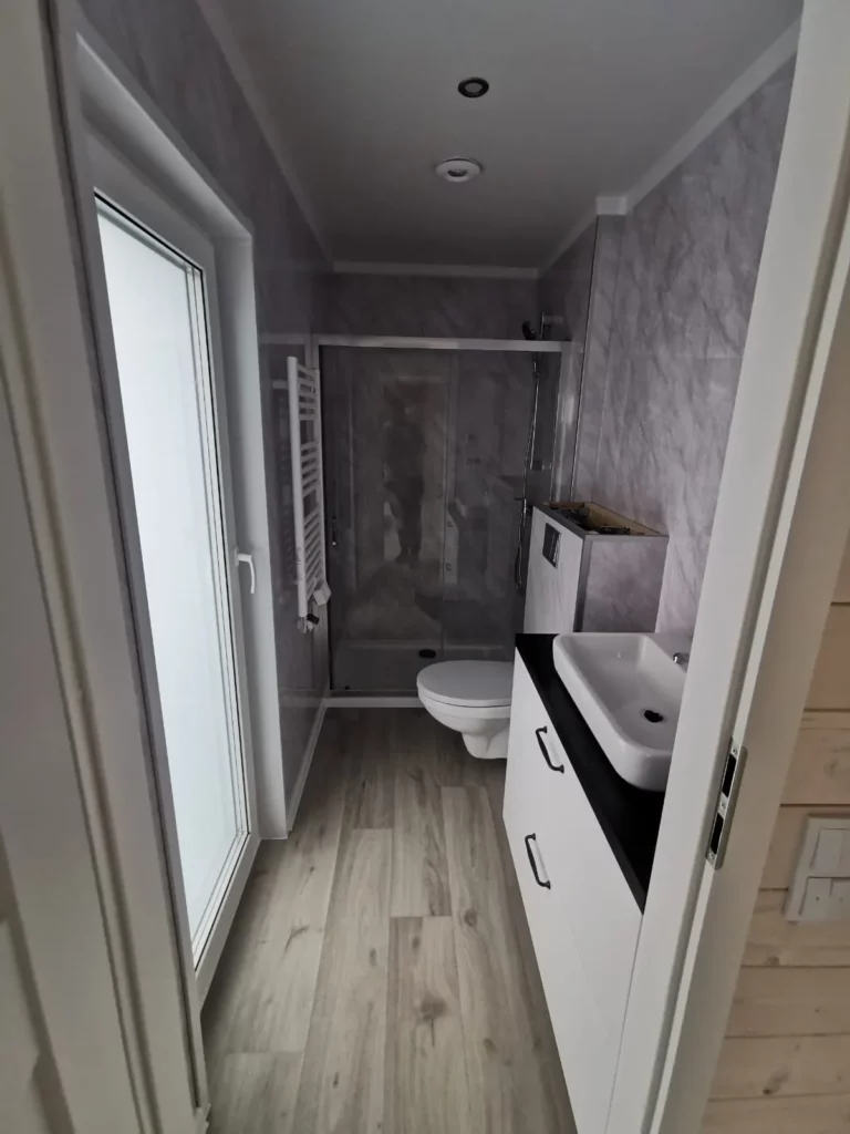 Badezimmer in einem winterfesten Mobilheim der MONO-Serie von Compact Haus mit grauer Wandverkleidung in Marmoroptik, Glasdusche, WC und weißem Waschtisch.