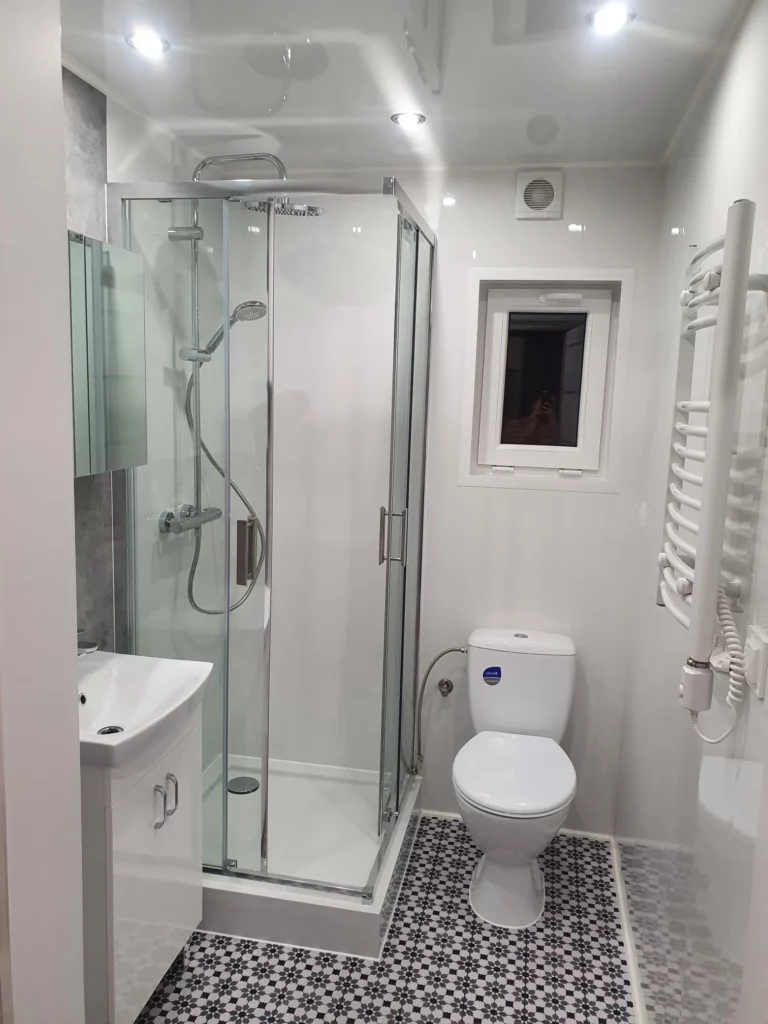 Helles, modernes Badezimmer in einem Compact Haus der MONO-Serie mit einer Eck-Glasdusche, gemusterten Mosaik-Bodenfliesen, WC, weißem Waschtisch und Handtuchheizkörper.