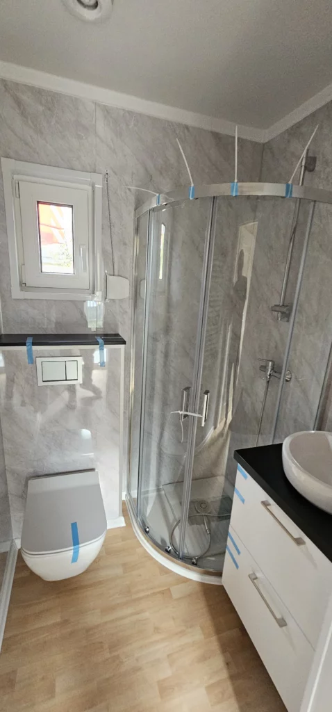 Modernes Badezimmer in einem Mobilheim der MONO-Serie von Compact Haus mit einer gläsernen Runddusche, Wand-WC, Waschtisch mit Aufsatzwaschbecken und Wänden in heller Marmoroptik.