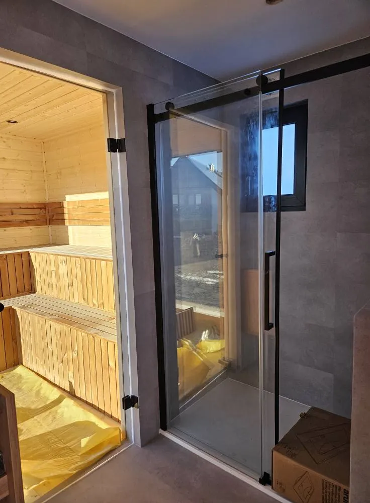 Blick in das moderne Badezimmer eines Compact Hauses der MONO-Serie mit direktem Zugang zu einer integrierten Holzsauna und einer bodengleichen Glasdusche mit schwarzem Rahmen.