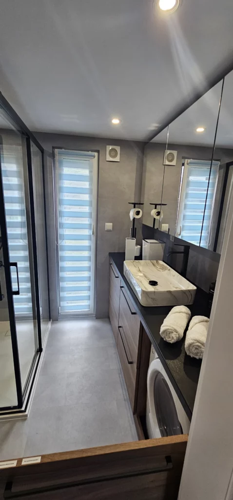 Luxuriöses Badezimmer im Compact Haus der MONO-Serie mit mattschwarzer Regendusche, großem Waschtisch in Holzoptik, Aufsetzwaschbecken in Marmoroptik und hinterleuchtetem Spiegel.