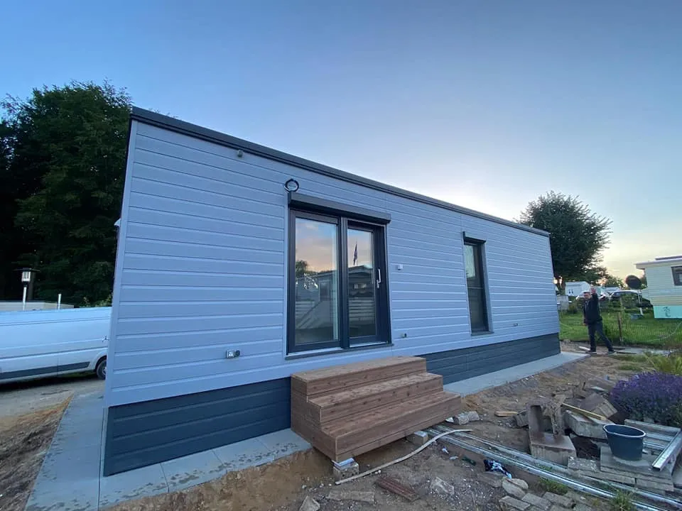 Modernes winterfestes Mobilheim der MONO-Serie von Compact Haus mit hellgrauer Außenfassade, anthrazitfarbenem Sockel und einer hölzernen Eingangstreppe.