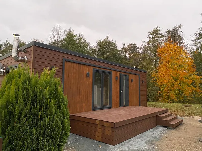 Modernes Mobilheim von Compact Haus mit Flachdach, Edelstahlschornstein, Holzterrasse und dunkelbrauner Holzfassade.