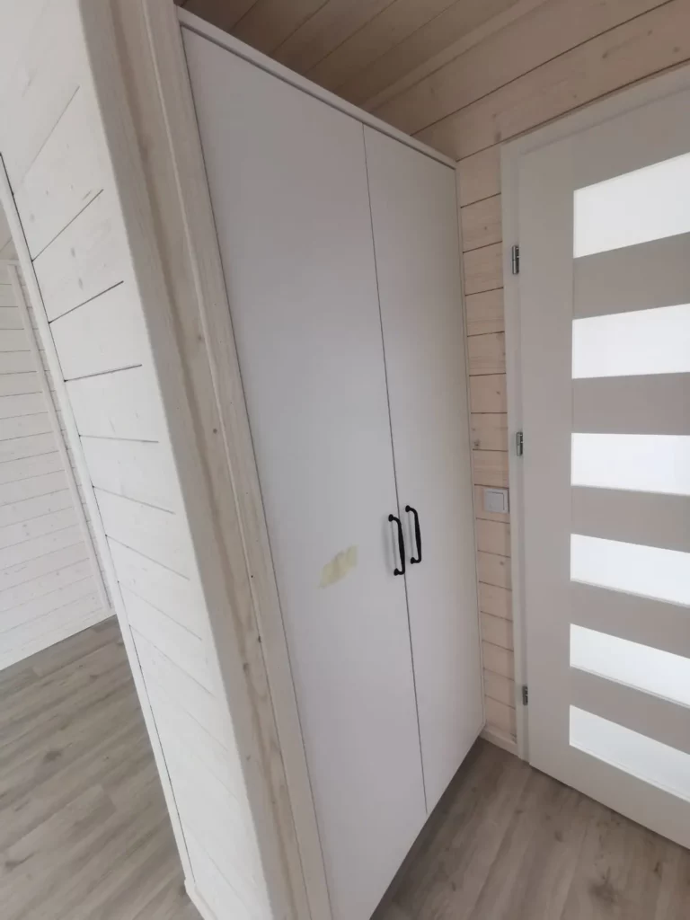 Eingangsbereich eines Compact Hauses der MONO-Serie mit hellem Holzboden, weißer Echtholz-Wandverkleidung, einer modernen Innentür und einem passgenauen weißen Einbauschrank mit schwarzen Griffen.