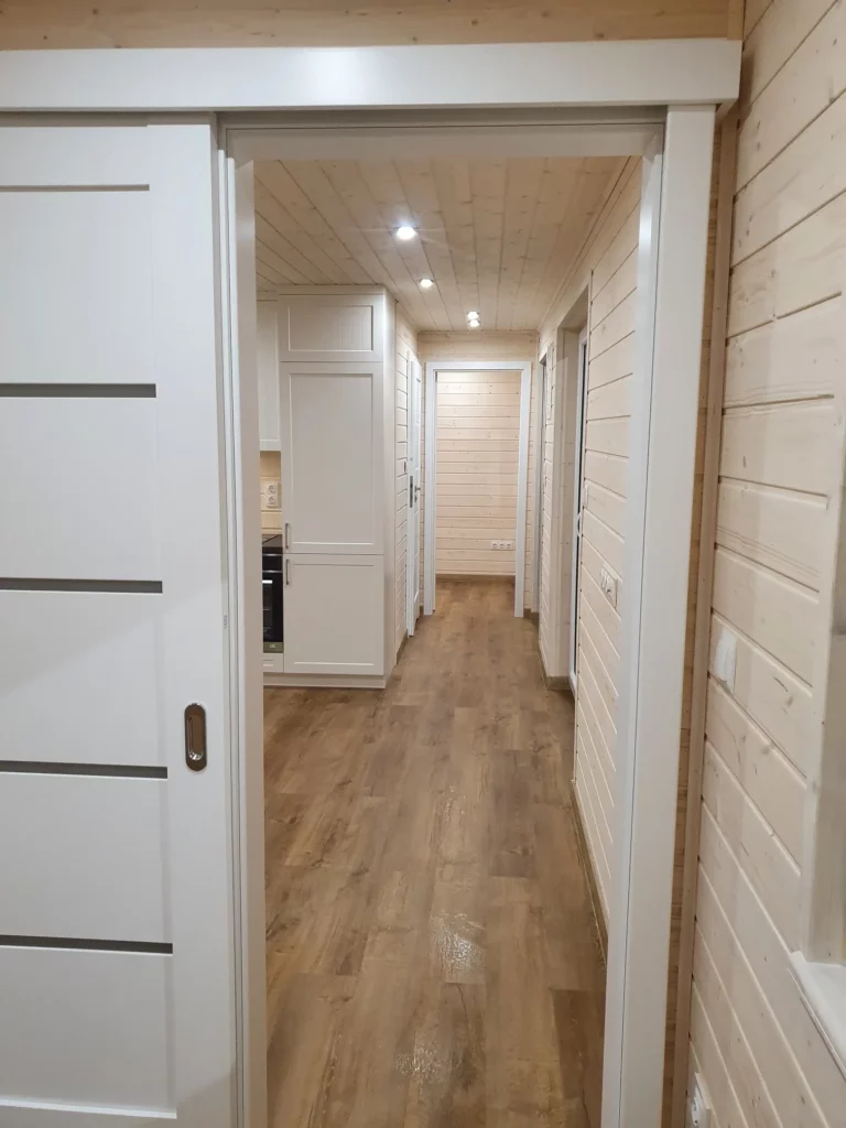 Blick durch eine geöffnete weiße Schiebetür in den langen Flur eines Compact Hauses der MONO-Serie mit hellen Echtholzwänden, LED-Deckenspots und dunklem Vinylboden.