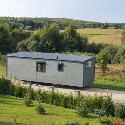 Elegantes Badezimmer im Mobilheim der MONO-Serie von Compact Haus mit bodengleicher Glasdusche, Waschtischunterschrank aus Holzlamellen, beleuchtetem Spiegelschrank und moderner Wandverkleidung in Betonoptik.