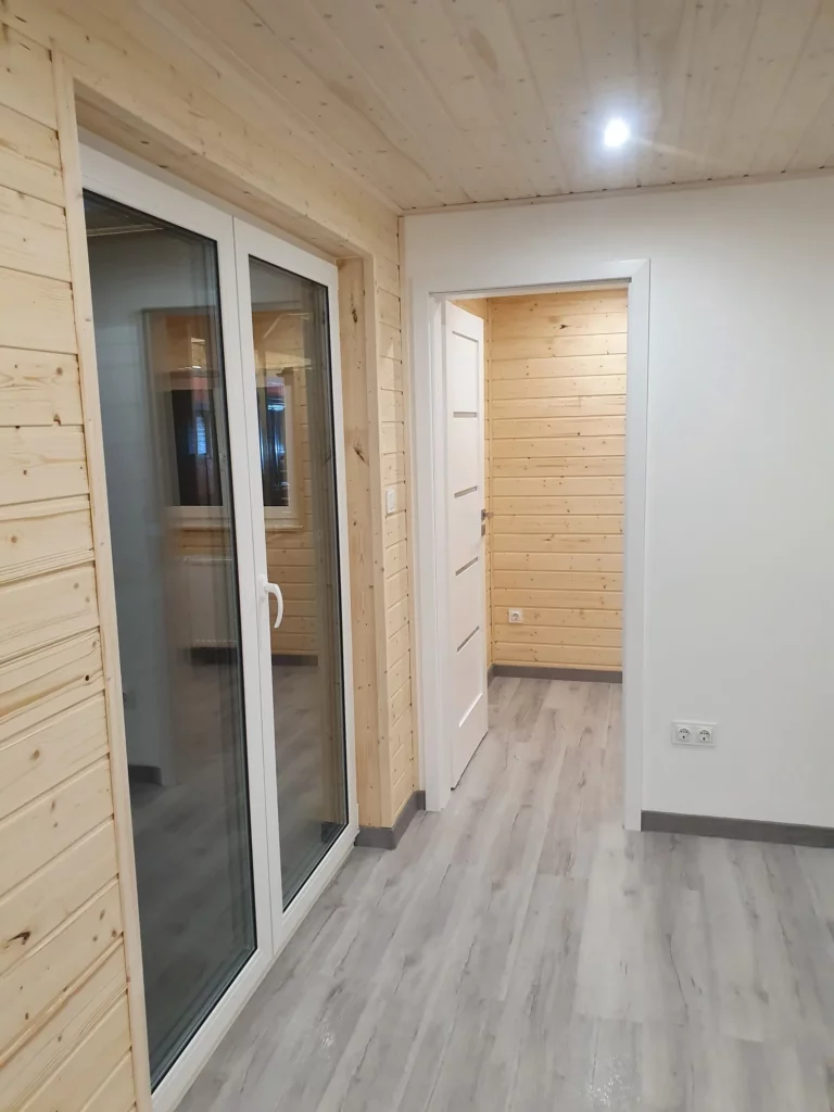 Lichtdurchfluteter Innenraum im Mobilheim der MONO-Serie von Compact Haus mit hellen Echtholzwänden, weißer Innentür, moderner Terrassentür und grauem Laminatboden.