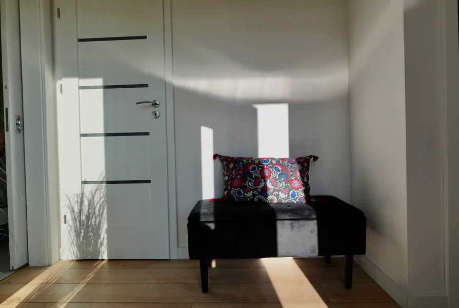Stilvolle Garderoben-Sitzbank mit gemustertem Kissen vor einer glatten weißen Innenwand im sonnendurchfluteten Flur eines Compact Haus Mobilheims.