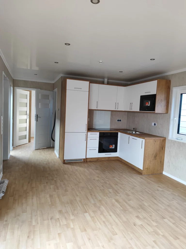 Moderne Einbauküche in L-Form mit weißen Fronten und Korpus in Holzoptik im unmöblierten Innenraum eines Compact Hauses der MONO-Serie mit hellem Vinylboden.
