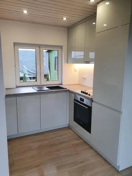 Kompakte L-Küche in einem Mobilheim der MONO-Serie von Compact Haus mit hellgrauen Hochglanzfronten, einer Arbeitsplatte in Holzoptik und einem großen Küchenfenster.