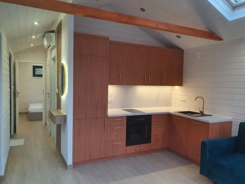 Moderne L-Küche in einem Compact Haus der MONO-Serie mit warmen Holzfronten, weißer Arbeitsplatte, goldenen Griffen und einer passenden goldenen Spültischarmatur.