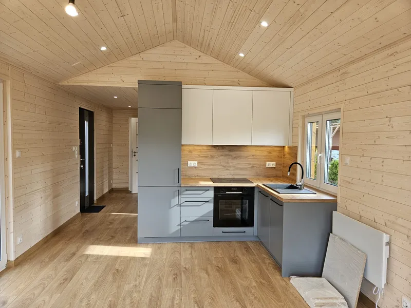 Kompakte graue Einbauküche mit weißen Oberschränken in einem Compact Haus der MONO-Serie inmitten von hellen, skandinavischen Holzwänden und Holzdecken.