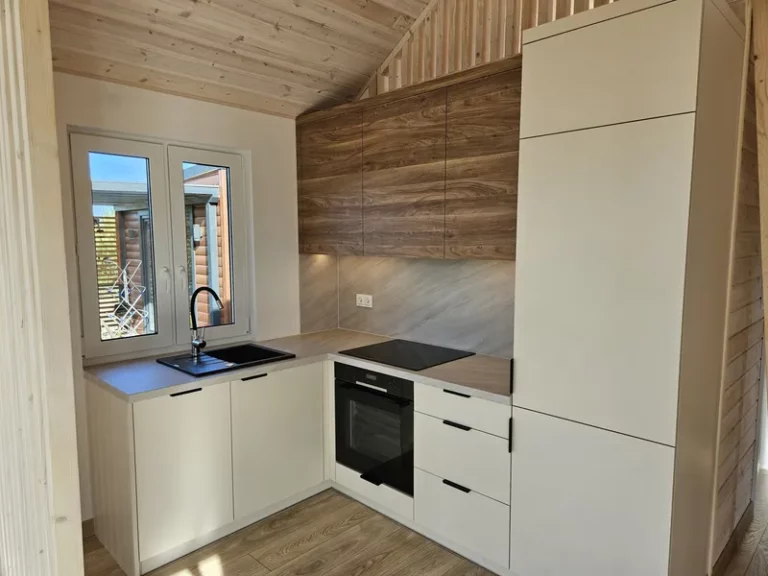 Eingepasste L-Küche in einem Compact Haus der MONO-Serie mit cremeweißen Fronten, Oberschränken in warmer Holzoptik und einer passenden Arbeitsplatte unter einer schrägen Holzdecke.