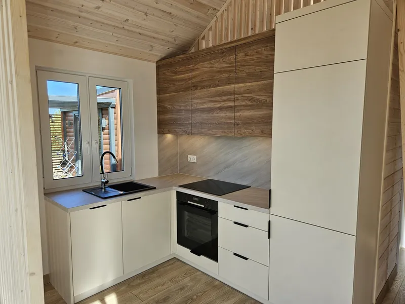 Eingepasste L-Küche in einem Compact Haus der MONO-Serie mit cremeweißen Fronten, Oberschränken in warmer Holzoptik und einer passenden Arbeitsplatte unter einer schrägen Holzdecke.