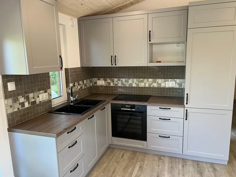 Moderne Einbauküche im Landhausstil in einem Compact Haus der MONO-Serie mit hellgrauen Rahmenfronten, dunkler Holzarbeitsplatte und einem Fliesenspiegel in Mosaikoptik.