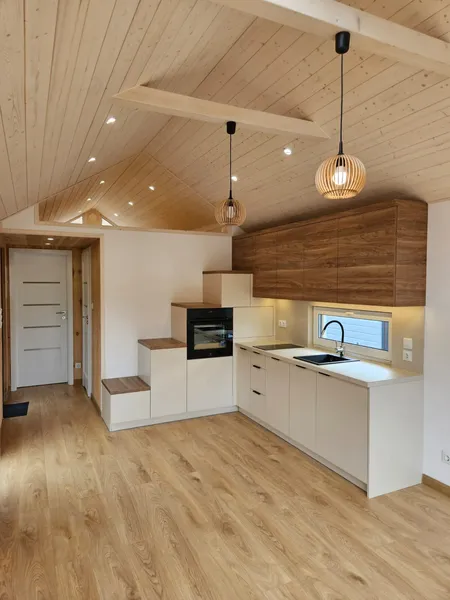 Moderne Einbauküche in einem Compact Haus der MONO-Serie mit integrierter Treppe zum Loft, hölzernen Oberschränken, hellem Echtholzboden und stilvollen Pendelleuchten unter einer hohen Holzdecke.