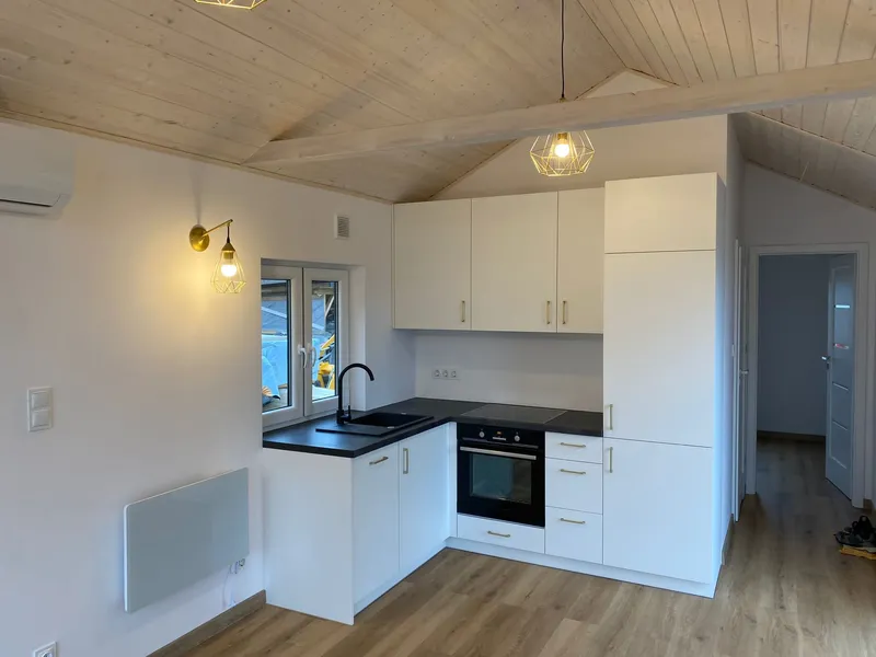 Minimalistische weiße Einbauküche in L-Form mit schwarzer Arbeitsplatte, goldfarbenen Drahtleuchten und einer hohen Decke in Holzoptik in einem Compact Haus der MONO-Serie.