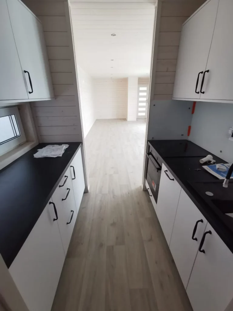 Zweizeilige Einbauküche in einem Compact Haus der MONO-Serie mit weißen Fronten, dunkler Arbeitsplatte, Kochfeld und Blick in den angrenzenden Wohnbereich.