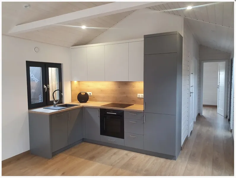 Moderne L-Küche in einem Compact Haus der MONO-Serie mit anthrazitgrauen Unterschränken, weißen Oberschränken, einer Nischenrückwand in Holzoptik und LED-Unterbaubeleuchtung.