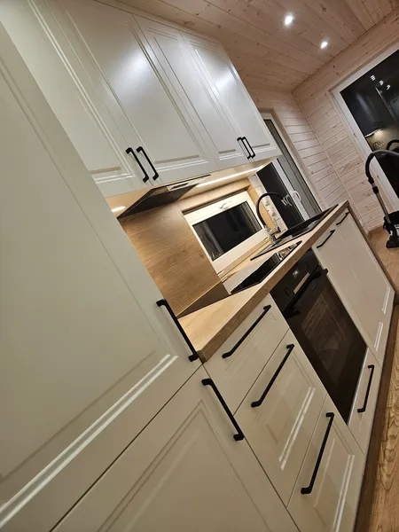 Elegante weiße Landhausküche in einem Mobilheim der MONO-Serie von Compact Haus mit schwarzen Griffen, einer Arbeitsplatte in warmer Holzoptik und LED-Nischenbeleuchtung.