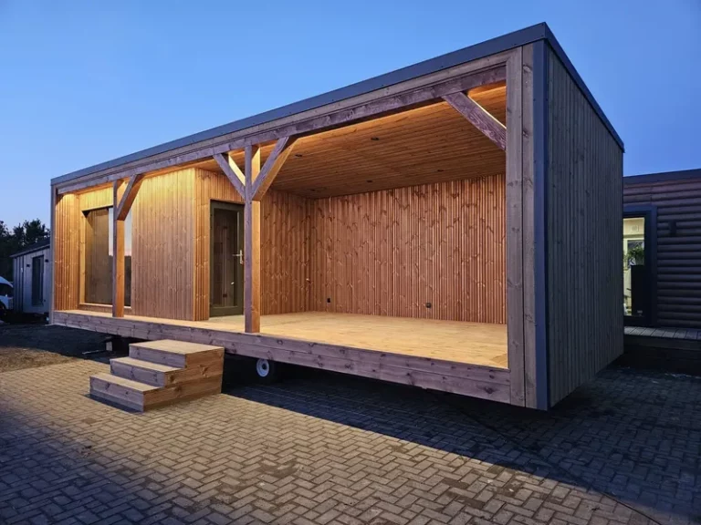 Schräge Außenansicht eines winterfesten Compact Hauses der MONO-Serie mit einer großzügigen, überdachten Holzterrasse, moderner LED-Deckenbeleuchtung und der Eingangstür zur Sauna.