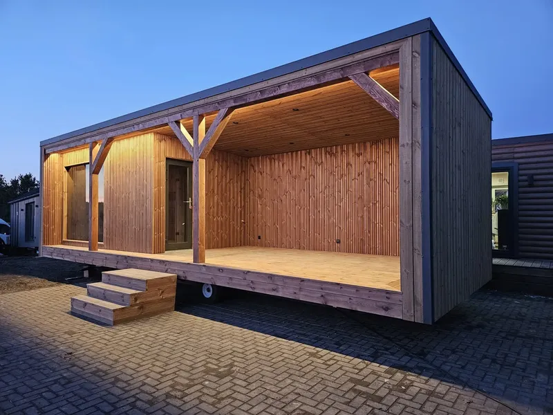 Schräge Außenansicht eines winterfesten Compact Hauses der MONO-Serie mit einer großzügigen, überdachten Holzterrasse, moderner LED-Deckenbeleuchtung und der Eingangstür zur Sauna.