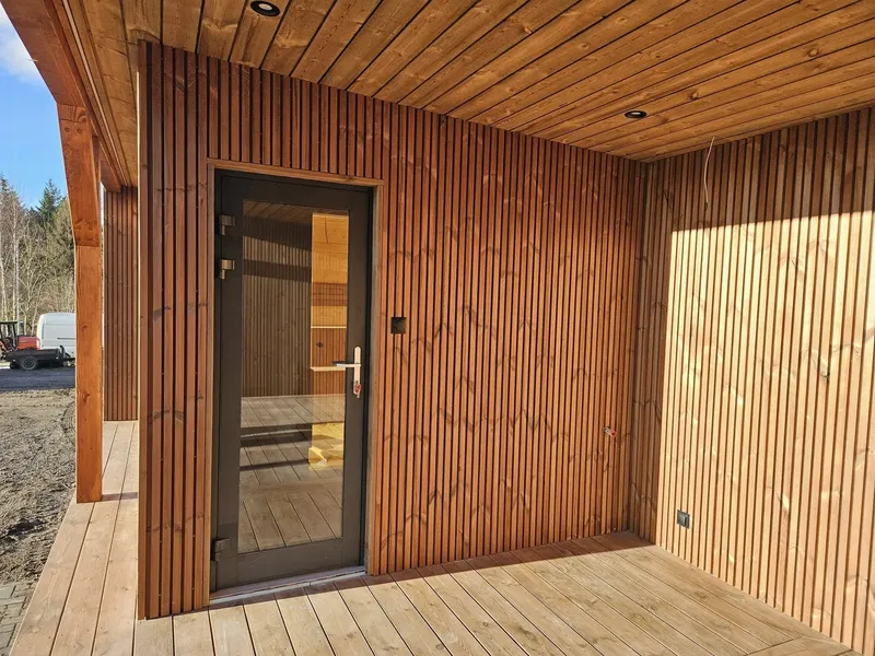 Detailansicht der verglasten Saunatür auf der überdachten Terrasse eines Compact Hauses der MONO-Serie mit moderner Rombus-Holzverkleidung an den Wänden.