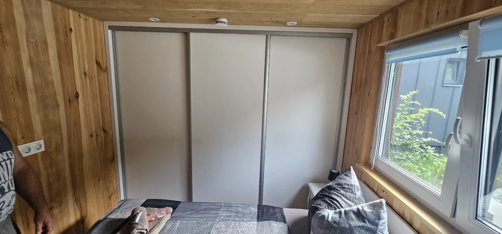 Gemütlicher Schlafraum in einem Compact Haus der MONO-Serie mit einem großen, dreitürigen Einbauschrank mit Schiebetüren, Echtholzwänden und integrierten LED-Deckenstrahlern.