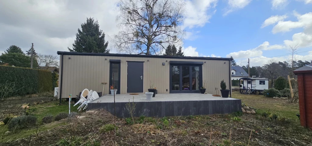Winterfestes Mobilheim der MONO-Serie von Compact Haus mit hellbeiger Fassade, anthrazitfarbener Eingangstür, großen Fensterelementen und einer davor liegenden Terrasse im Garten.