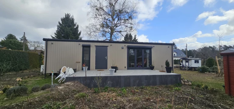 Winterfestes Mobilheim der MONO-Serie von Compact Haus mit hellbeiger Fassade, anthrazitfarbener Eingangstür, großen Fensterelementen und einer davor liegenden Terrasse im Garten.