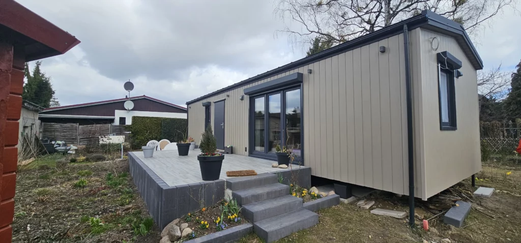 Perspektivische Ansicht eines winterfesten Compact Hauses der MONO-Serie mit hellbeiger Holzfassade, großer Holzterrasse, grauen Blockstufen aus Beton und bepflanzten Blumentöpfen.